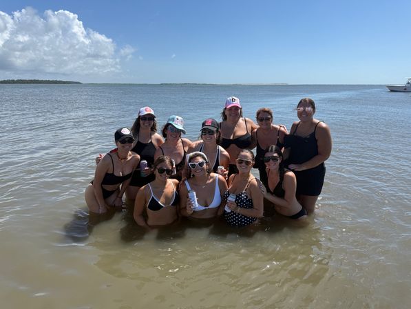 Group of friends waist-deep in shallow coastal water on a sunny beach day, wearing swimsuits, hats and sunglasses, smiling and holding canned drinks with a calm bay, distant shoreline and a boat on the horizon.
