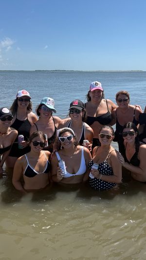 Smiling group of women in swimsuits standing waist-deep in shallow coastal bay water, holding drinks with distant islands and a boat under a bright blue sky