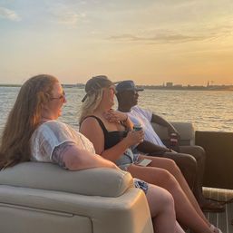 Three friends laughing on a pontoon boat at sunset, sipping drinks on a calm waterfront cruise with a distant skyline.