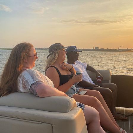 Three friends laughing on a pontoon boat at sunset, sipping drinks on a calm waterfront cruise with a distant skyline.