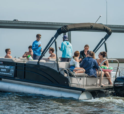 Group of friends laughing and relaxing on a pontoon party boat cruising under a highway bridge on calm water, sunny day