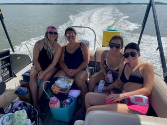 Four friends in swimsuits smiling and holding drinks on a pontoon boat with a cooler of snacks and towels, wake trailing across a sunny lake.