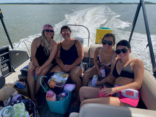 Four friends in swimsuits smiling and holding drinks on a pontoon boat with a cooler of snacks and towels, wake trailing across a sunny lake.