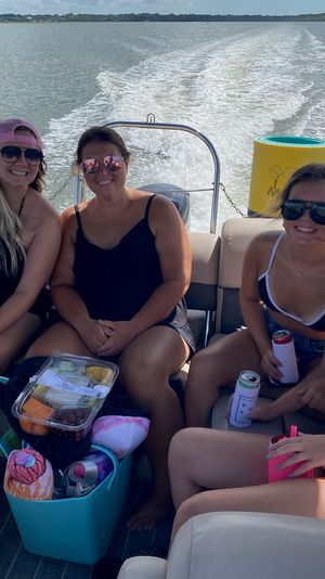 Four friends in swimsuits and sunglasses smiling on a pontoon boat, holding drinks and snacks with a foamy wake trailing across a marshy bay under a sunny sky.