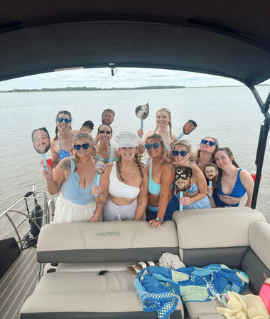 Bachelorette boat party group photo on calm water — smiling women in swimsuits and sunglasses under a canopy, holding novelty face-on-stick props with a distant tree-lined shoreline in the background.
