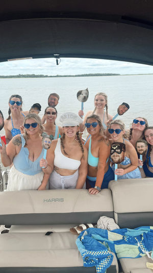 Bachelorette boat party group photo on calm water — smiling women in swimsuits and sunglasses under a canopy, holding novelty face-on-stick props with a distant tree-lined shoreline in the background.