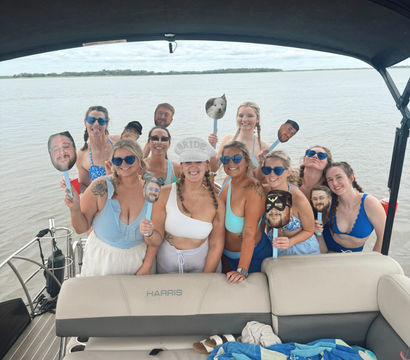Bachelorette boat party group photo on calm water — smiling women in swimsuits and sunglasses under a canopy, holding novelty face-on-stick props with a distant tree-lined shoreline in the background.
