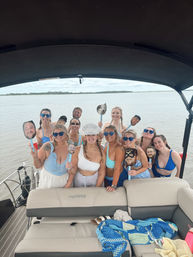Group of friends in swimsuits on a pontoon boat on a calm lake, smiling and holding novelty face cutouts with the bride wearing a 'BRIDE' hat for a playful bachelorette party.