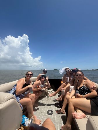 Group of friends relaxing on a pontoon boat on a sunny summer day, laughing and holding drinks with a distant shoreline, calm water and large cumulus clouds under a clear blue sky.