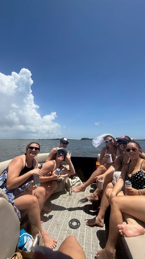 Group of friends relaxing on a pontoon boat on a sunny summer day, laughing and holding drinks with a distant shoreline, calm water and large cumulus clouds under a clear blue sky.