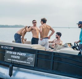 Group of shirtless young men socializing and lounging on a party pontoon boat on a calm North Carolina lake, one man at the helm.