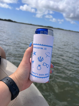 Hand holding a slim can in a white-and-blue Charleston, SC personalized can cooler on a boat over calm marsh waters with blue sky and clouds