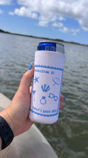Hand holding a slim can in a white-and-blue Charleston, SC personalized can cooler on a boat over calm marsh waters with blue sky and clouds