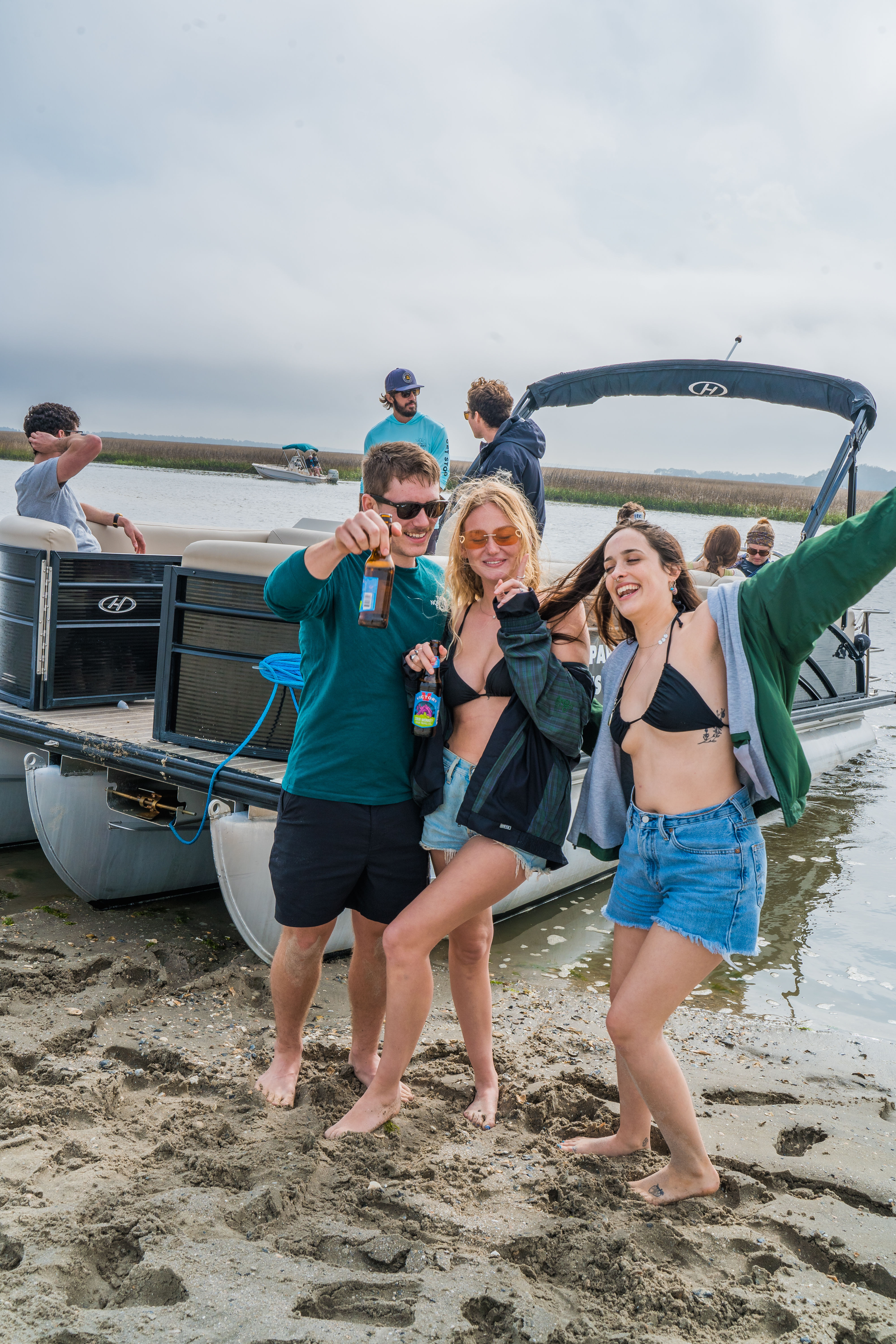 Three friends laughing on a sandy coastal inlet beside a pontoon boat, wearing swimwear and holding beers with marsh grasses and other boats in the background.