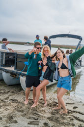 Three friends laughing on a sandy coastal inlet beside a pontoon boat, wearing swimwear and holding beers with marsh grasses and other boats in the background.