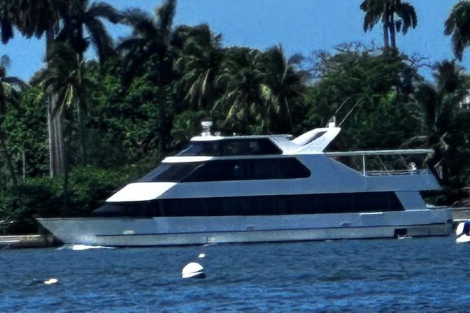 White luxury yacht moored by buoys in blue water off a palm-lined tropical shoreline under a clear sky