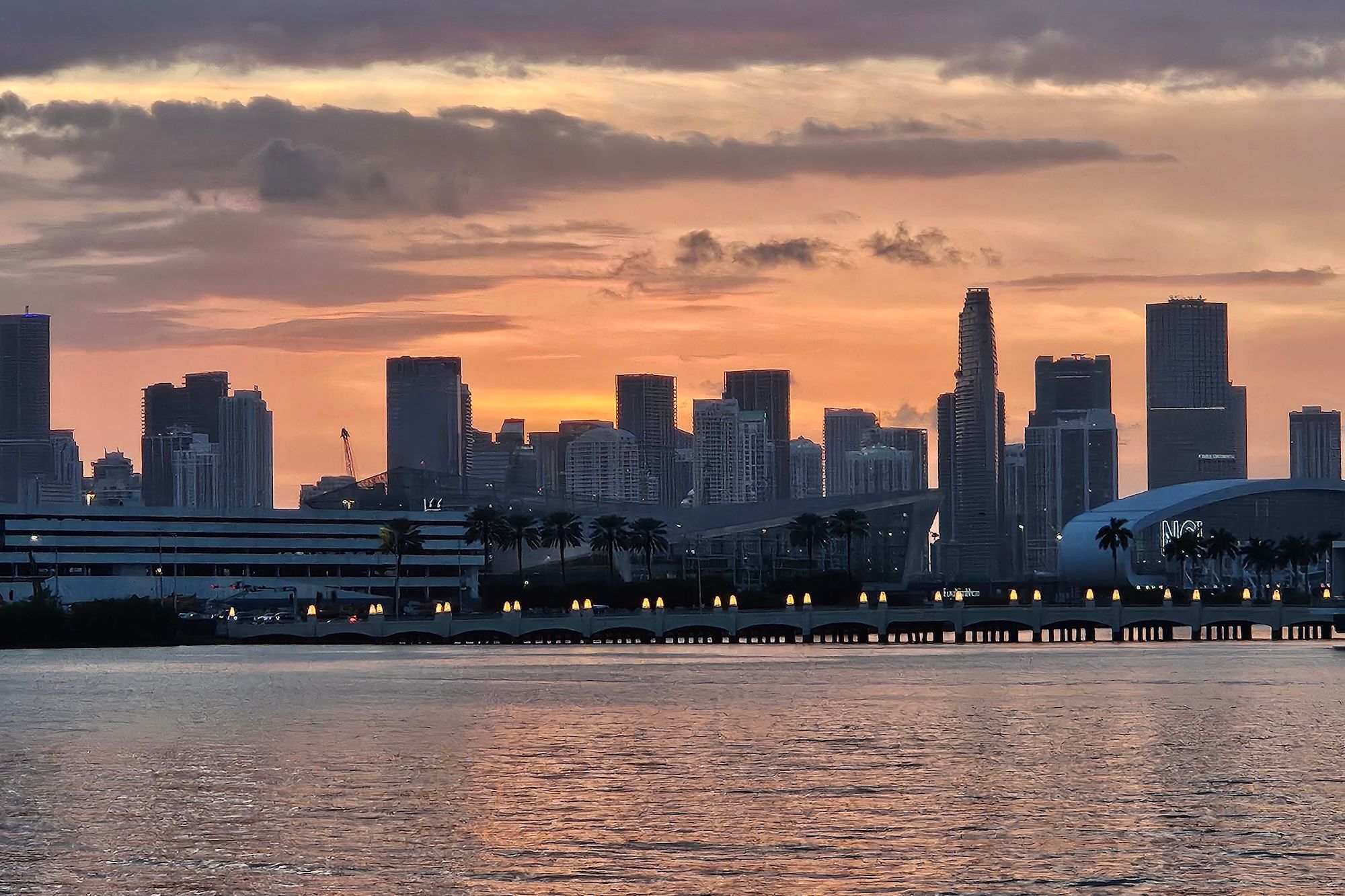 Miami skyline silhouette at sunset over Biscayne Bay, glowing orange sky, palm trees and lit bridge arches reflected on calm water.