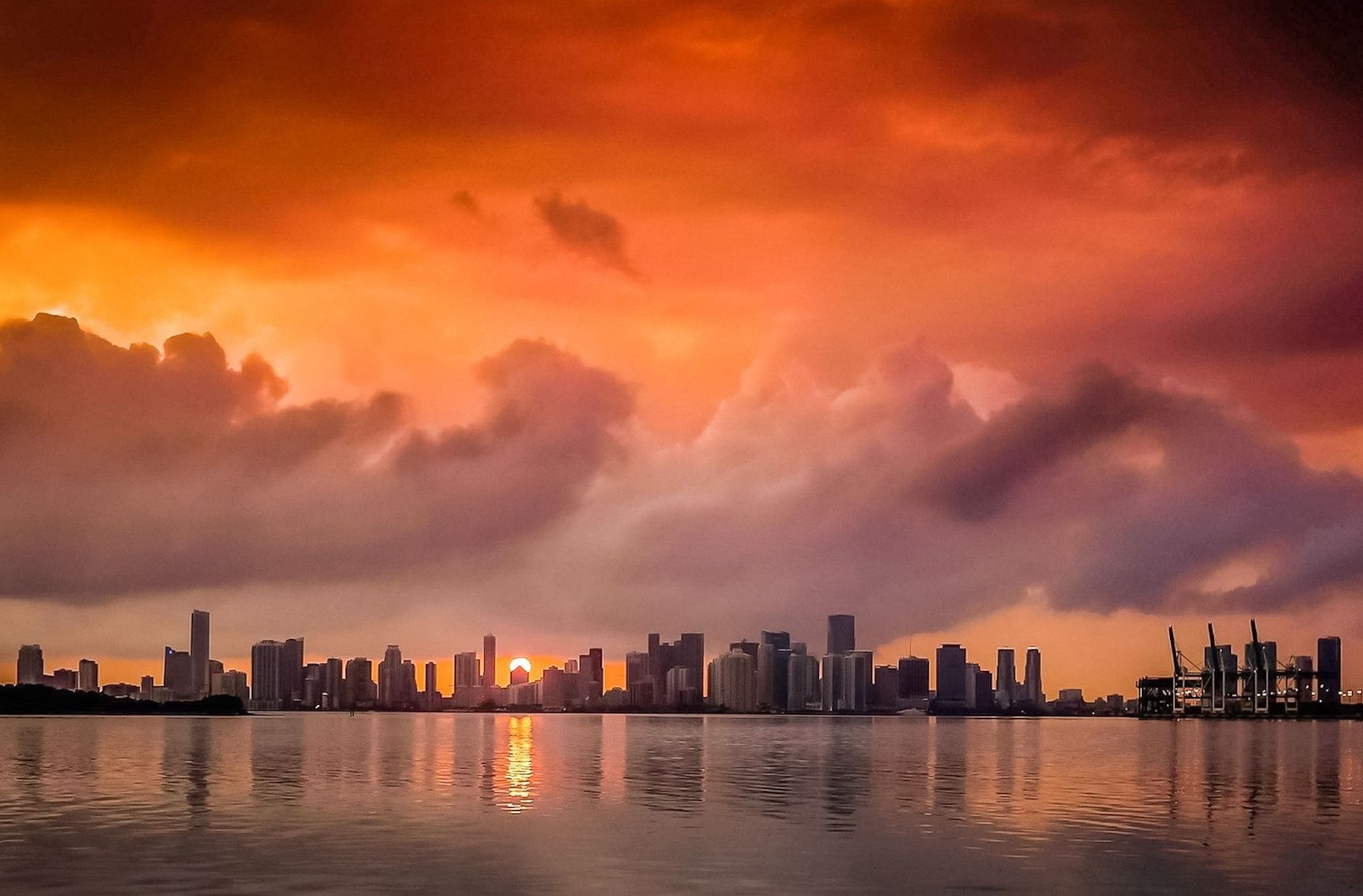 Fiery orange sunset over a coastal city skyline, dramatic clouds above high-rise buildings and port cranes reflected in calm bay waters.