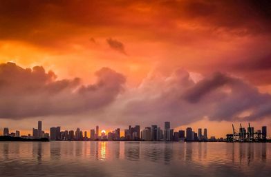 Fiery orange sunset over a coastal city skyline, dramatic clouds above high-rise buildings and port cranes reflected in calm bay waters.