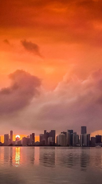 Fiery orange sunset over a coastal city skyline, dramatic clouds above high-rise buildings and port cranes reflected in calm bay waters.