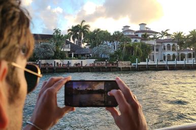 Person on a boat snapping a smartphone photo of a palm-lined waterfront mansion at golden hour