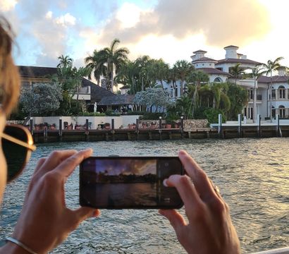 Person on a boat snapping a smartphone photo of a palm-lined waterfront mansion at golden hour