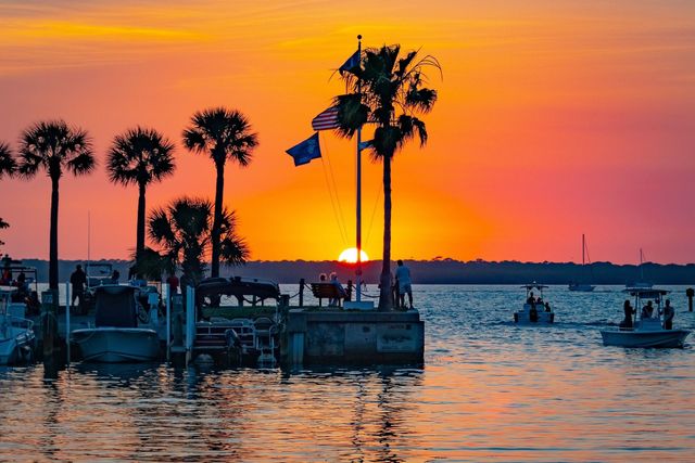 Golden-orange coastal sunset over a marina, silhouetted palm trees and a flagpole on a pier, boats bobbing on reflective water and people enjoying the view.