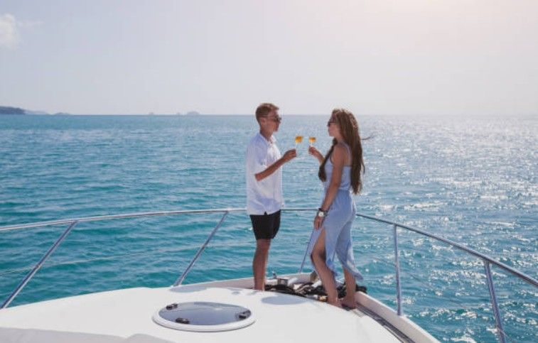 Couple toasting with drinks on the bow of a yacht over sparkling turquoise ocean on a sunny summer cruise.