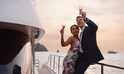 Romantic couple in dress and suit toasting with orange cocktails on a yacht deck at sunset, ocean and a distant island in view