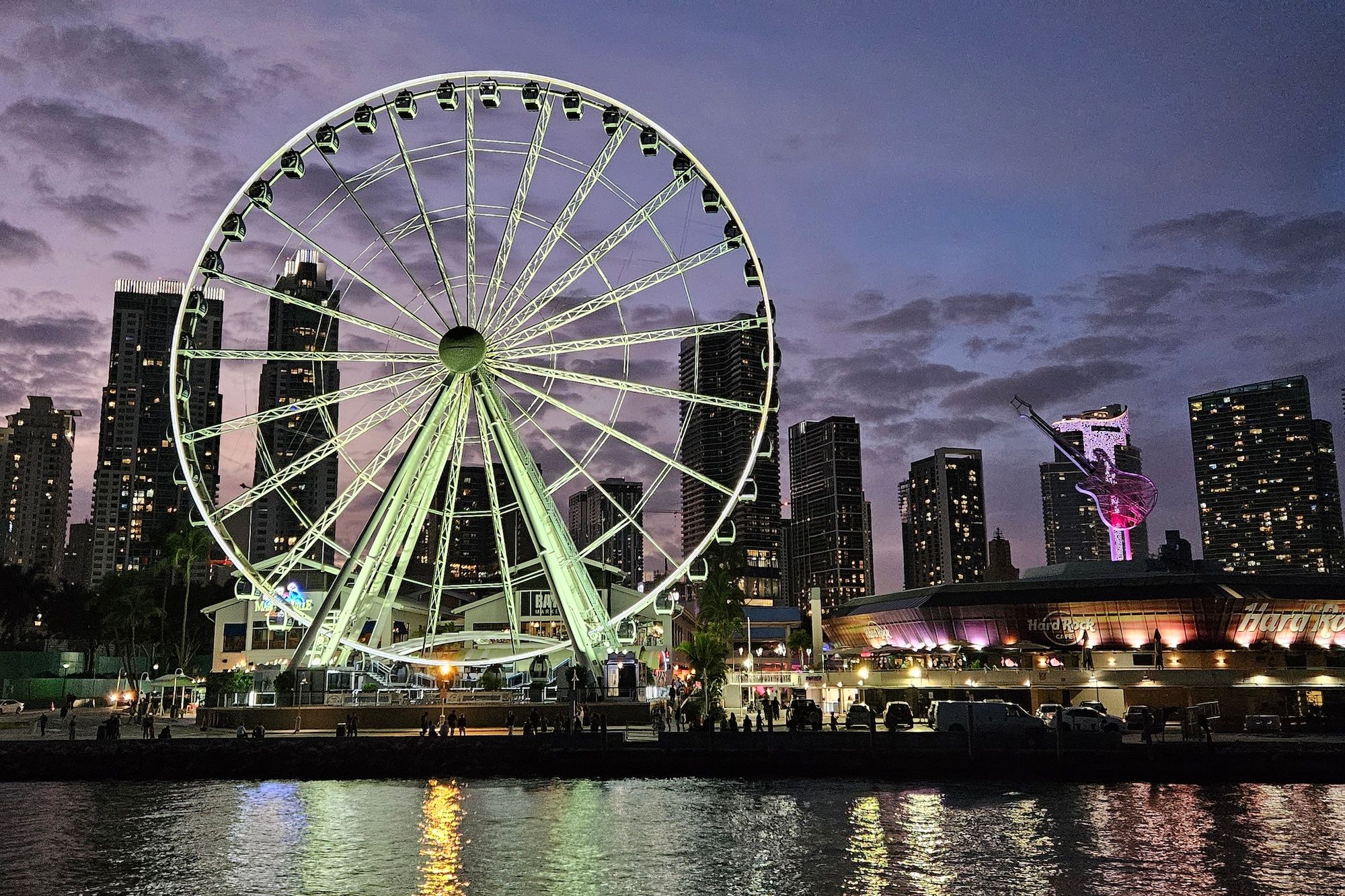 Illuminated Ferris wheel on an urban waterfront at dusk with a neon-lit guitar sculpture, high-rise skyline, and colorful reflections in the water.