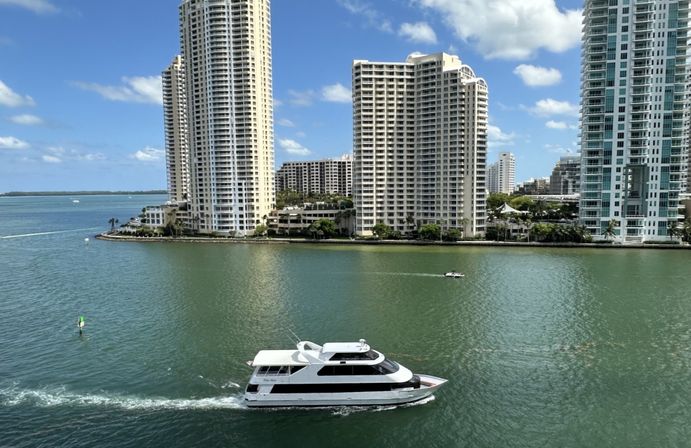 White luxury yacht cruising a turquoise bay past tall waterfront condominium towers under a bright blue sky.