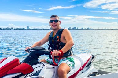 Smiling person in sunglasses and a life vest riding a red-and-white jet ski on calm coastal waters with a distant city skyline and bright blue sky.