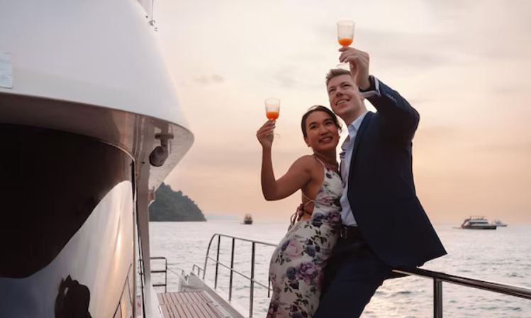 Couple on a yacht deck toasting with drinks at sunset over a calm ocean with a distant island, romantic sunset cruise