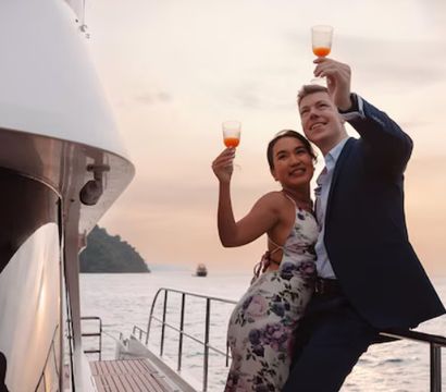 Couple on a yacht deck toasting with drinks at sunset over a calm ocean with a distant island, romantic sunset cruise