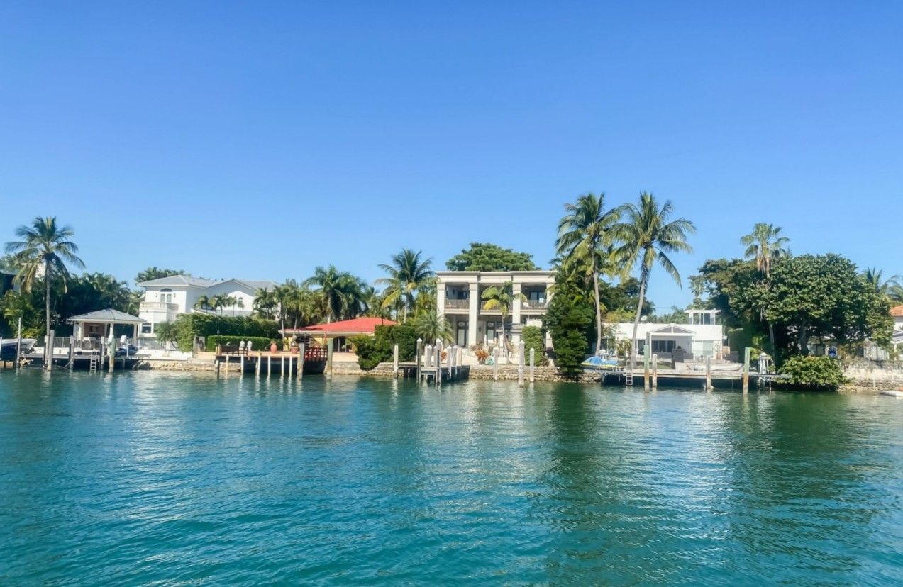 Sunny tropical waterfront with luxury homes, swaying palm trees and private docks along a turquoise coastal canal under a clear blue sky
