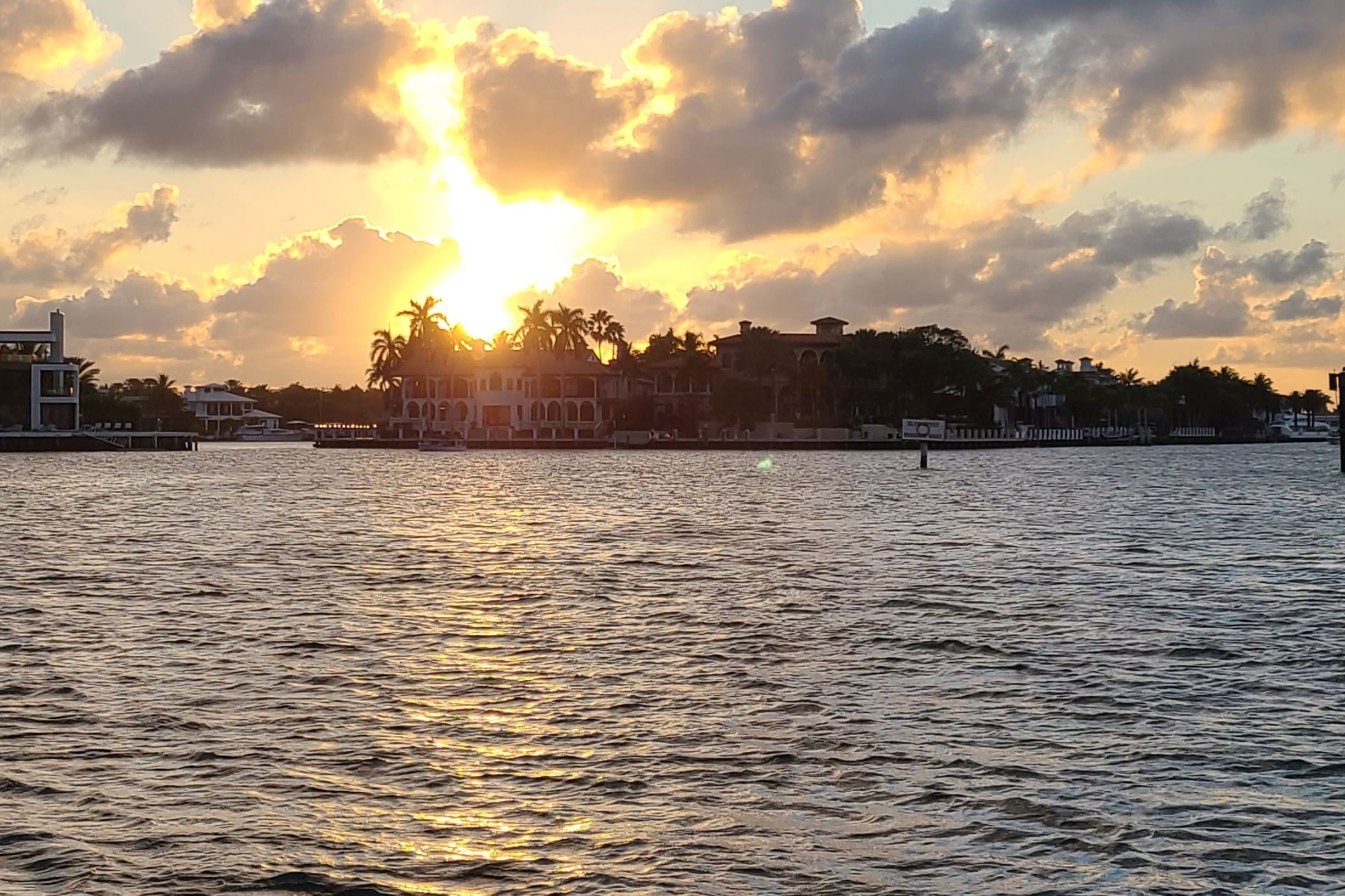 Golden sunset over a palm-lined waterfront with luxury homes, sunburst through clouds and shimmering reflections on rippled bay water.