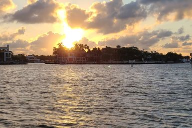 Golden sunset over a palm-lined waterfront with luxury homes, sunburst through clouds and shimmering reflections on rippled bay water.