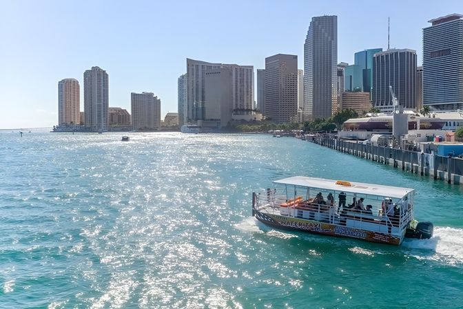 Water taxi with passengers cruising turquoise Biscayne Bay past the Miami high-rise skyline, sun sparkling on the water