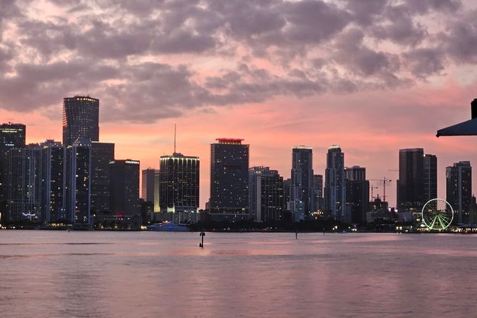 Miami skyline at sunset over a calm bay — pink-orange clouds, lit high-rise buildings silhouetted against the sky, and a glowing Ferris wheel on the waterfront.