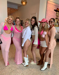 Six smiling women in pink and white beachwear, mesh cover-ups, cowboy boots and a pink cowgirl hat posing indoors at a boutique-style event.