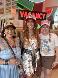 Three smiling women in boho-cowgirl outfits wearing decorated caps pose in a retro neon-lit interior in front of a 'VACANCY' sign — floral sundress, layered turquoise jewelry, studded belt, fringe bag, and casual graphic tee.