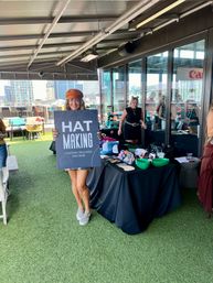 Woman on a downtown rooftop patio holding a sign reading “HAT MAKING” in front of a custom trucker hat bar table with colorful caps, patches and a city skyline in the background.