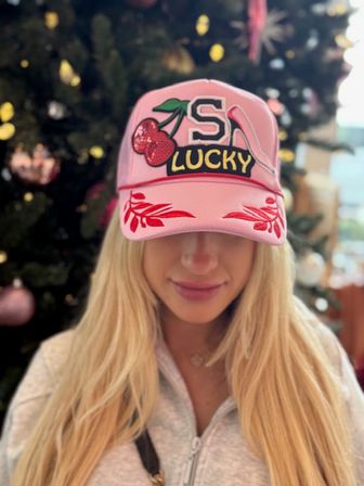 Person with long blonde hair wearing a pink baseball cap featuring a cherry patch, 'S' and "LUCKY" text and red embroidered brim, posed in front of a decorated Christmas tree.