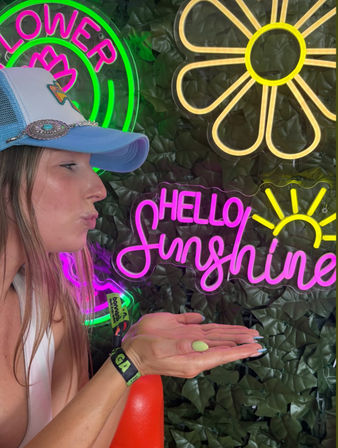 Playful profile of a woman in a light blue trucker hat blowing a kiss onto her cupped hand in front of a vibrant neon sign wall that reads "Hello Sunshine" with daisy and sun motifs