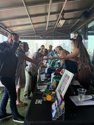 Rooftop market stall on an urban patio, shoppers exchanging goods with vendors over a table of colorful hats, patches and drinks with city skyline in the background.
