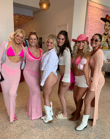 Six women posing in a bright boutique-style showroom wearing pink beachwear and crochet cover-ups, bikinis, cowboy boots and a pink cowboy hat, smiling in a fun group photo by artwork and pendant light.