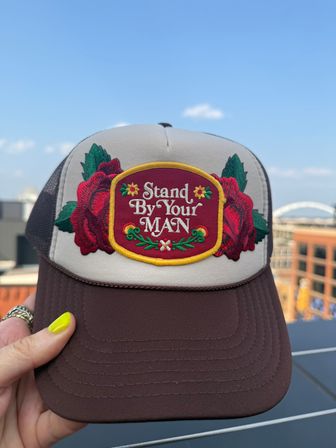 Vintage-style trucker hat with beige front and brown brim, embroidered red roses and yellow-trim patch reading 'Stand By Your Man', held by a hand with neon yellow nail polish against a blurred urban rooftop and blue sky.