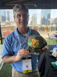 Smiling man in a blue polo holding a camo trucker hat with an embroidered tiger patch and “2016” numbers, wearing a VIP conference badge on an urban rooftop with a blurred city skyline