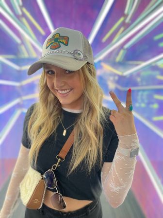 Smiling woman in a decorated baseball cap flashing a peace sign in a neon purple tunnel backdrop, wearing a cropped black tee over sheer long sleeves, layered necklaces, crossbody bag with sunglasses, and a turquoise ring.