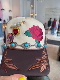 Beige and brown trucker hat with a bold embroidered red rose, turquoise concho chain, gold sunburst heart and pearl pins, and copper leaf-stitched brim, held by a hand with yellow nail polish against a blurred shop interior.